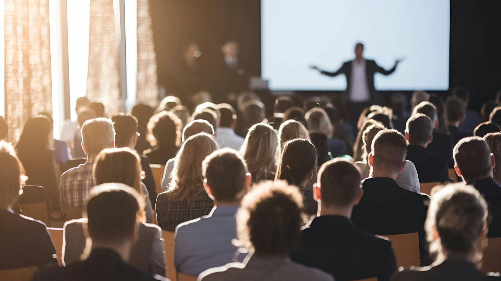 A speaker presents to a large audience in a sunlit auditorium, with a bright projection screen and professional atmosphere.