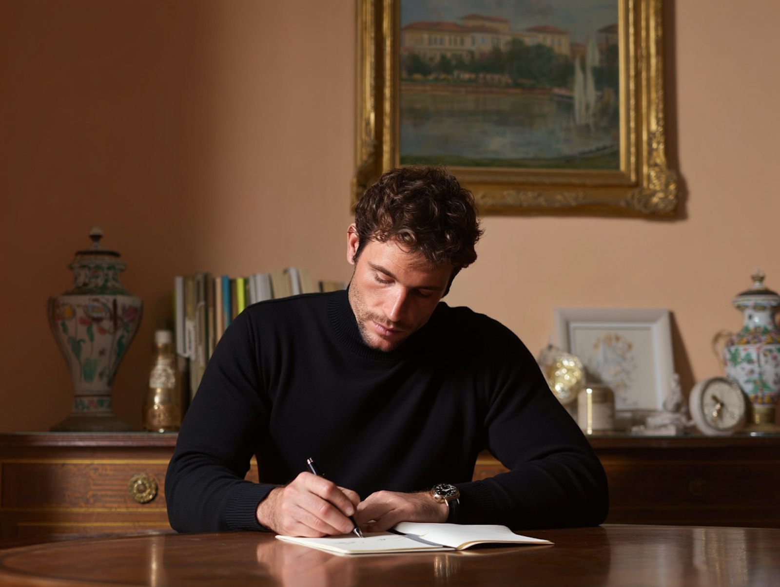 A man sits at a table writing in a warmly lit room, surrounded by elegant decor including vases, a painting, and books.